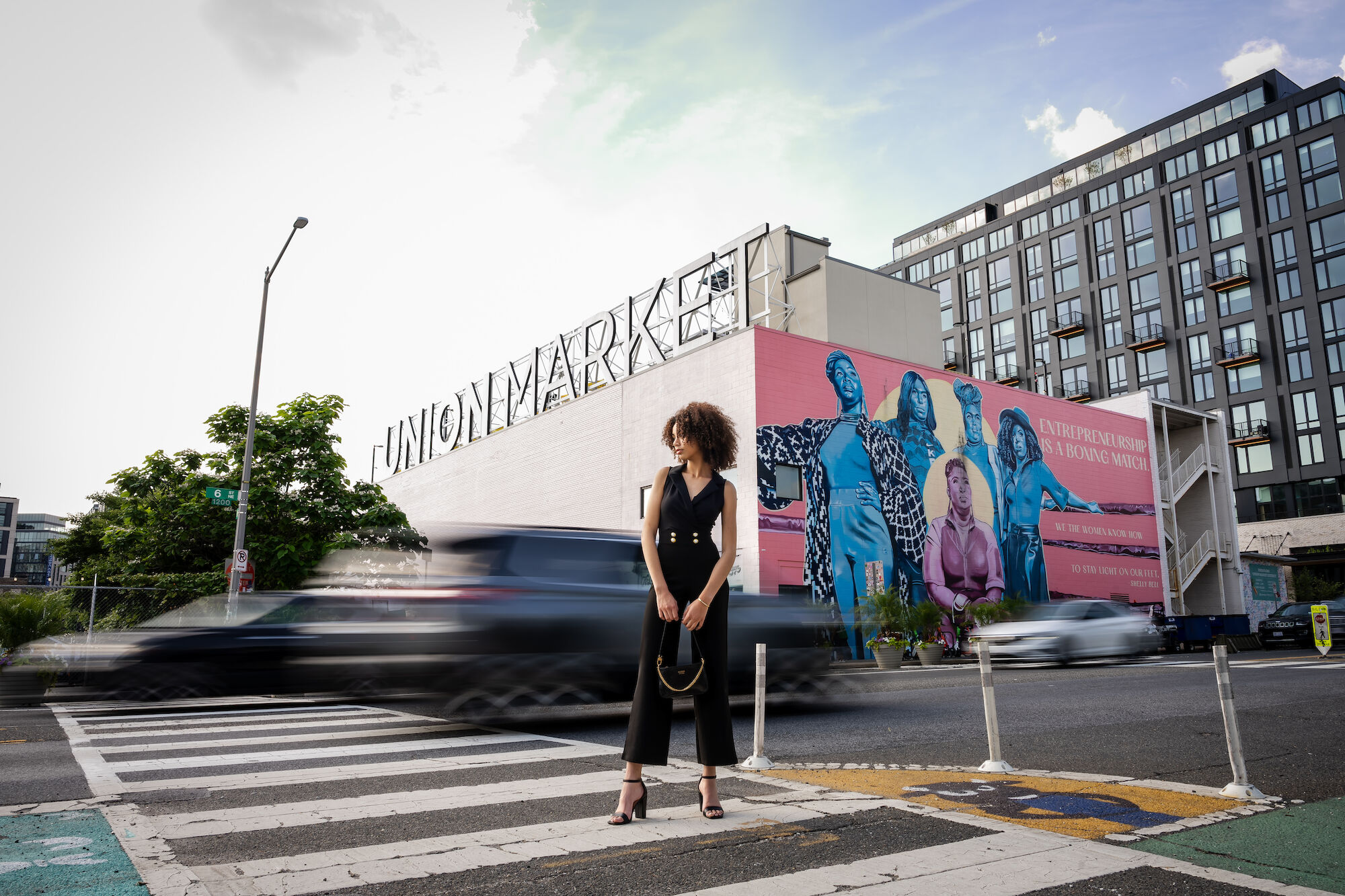 A person stands on a crosswalk in front of a mural and a Union Market building, while a car passes by.