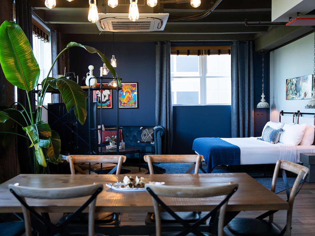 A modern room with a bed, dining area, large plant, wall art, and hanging lights, featuring a mix of wooden and industrial decor elements.