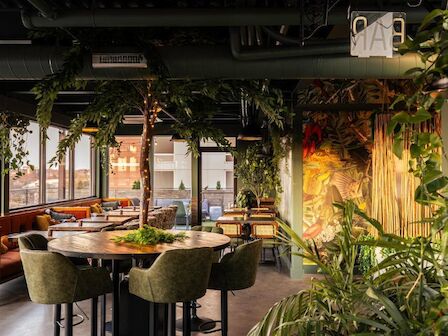 The image features a modern indoor restaurant with green decor, plants, and large windows that let in natural light, creating a refreshing atmosphere.