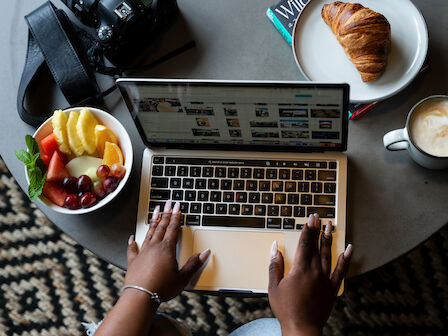 A person is using a laptop on a table with a bowl of fruit, a croissant on a plate, a cup of coffee, and a camera.