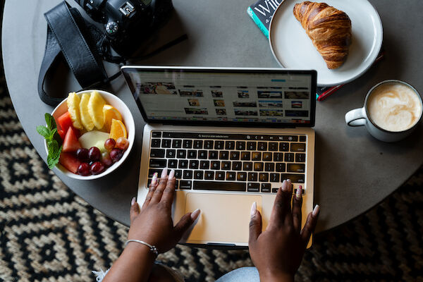 A person is using a laptop on a table with a bowl of fruit, a croissant on a plate, a cup of coffee, and a camera.