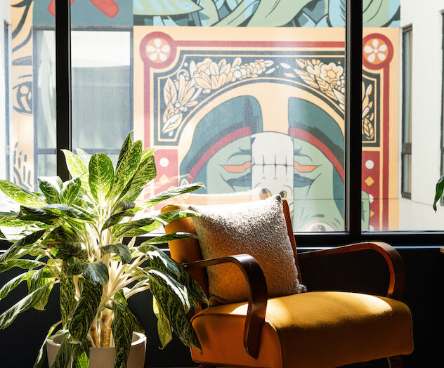 A cozy room with a yellow armchair, white pillow, potted plant, and a colorful mural outside the large window.