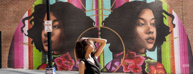 A person poses in front of a vibrant mural featuring two large portraits with colorful backgrounds on a brick wall.