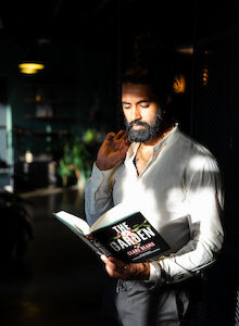 A bearded man in a white shirt is standing, reading a book titled 
