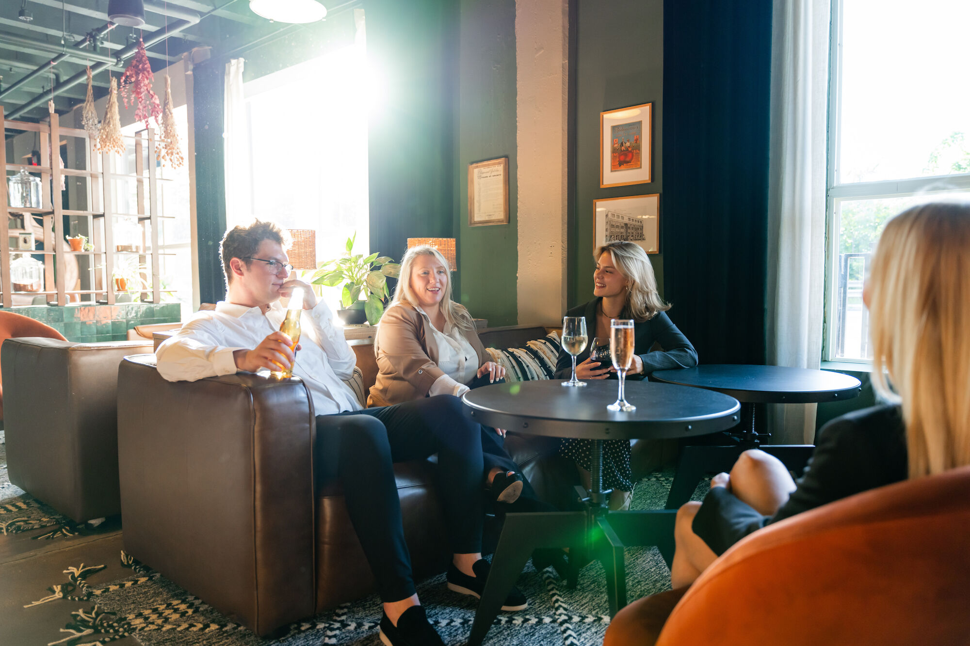 A group of people is sitting in a cozy, well-lit room, having a conversation over drinks, with modern decor and large windows in the background.