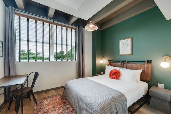 A cozy bedroom with a modern aesthetic features a bed with white sheets, a gray blanket, a red pillow, a wooden desk, and a large window.