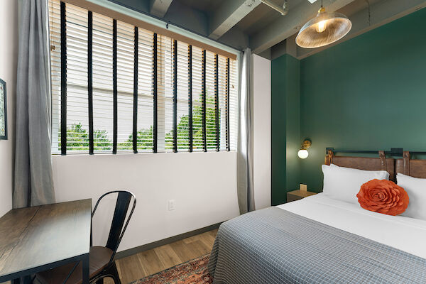 A small, modern bedroom with a bed, a desk, a chair, a window with blinds, a green wall, and a decorative pillow.