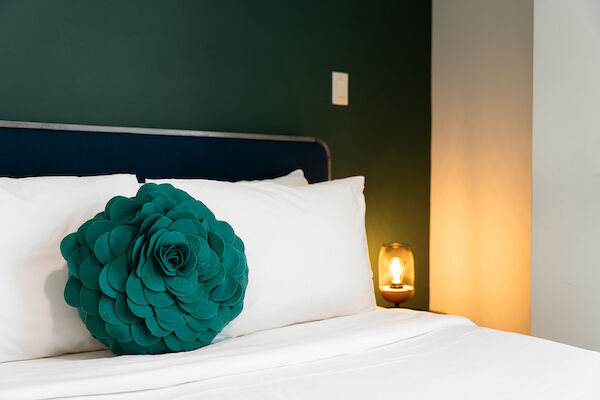 A neatly made bed with white linens features a green decorative pillow shaped like a flower and a lit bedside lamp on a dark background.