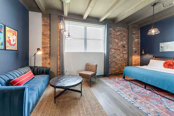 A modern bedroom with exposed brick walls, blue sofa, round coffee table, armchair, king-size bed, rug, art on walls, and hanging lights.