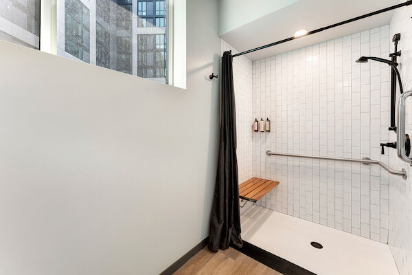 A modern shower area with white tiled walls, a wooden bench, black shower fixtures, and a grab bar. There is a curtain and a window nearby.