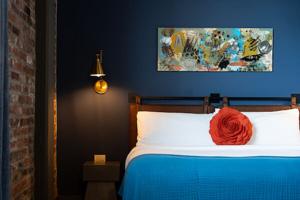 A neatly made bed with a blue blanket, a red flower cushion, brick wall, wall art, and a bedside table with a lamp in a cozy room.