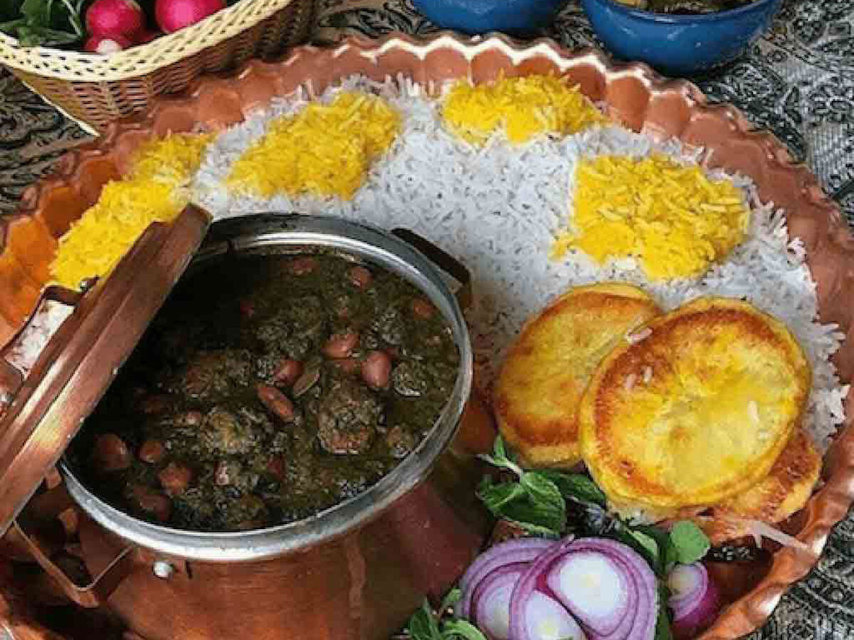 The image shows a traditional meal featuring rice topped with saffron, a pot of stew, sliced onions, herbs, radishes, yogurt, and another dish in blue bowls.