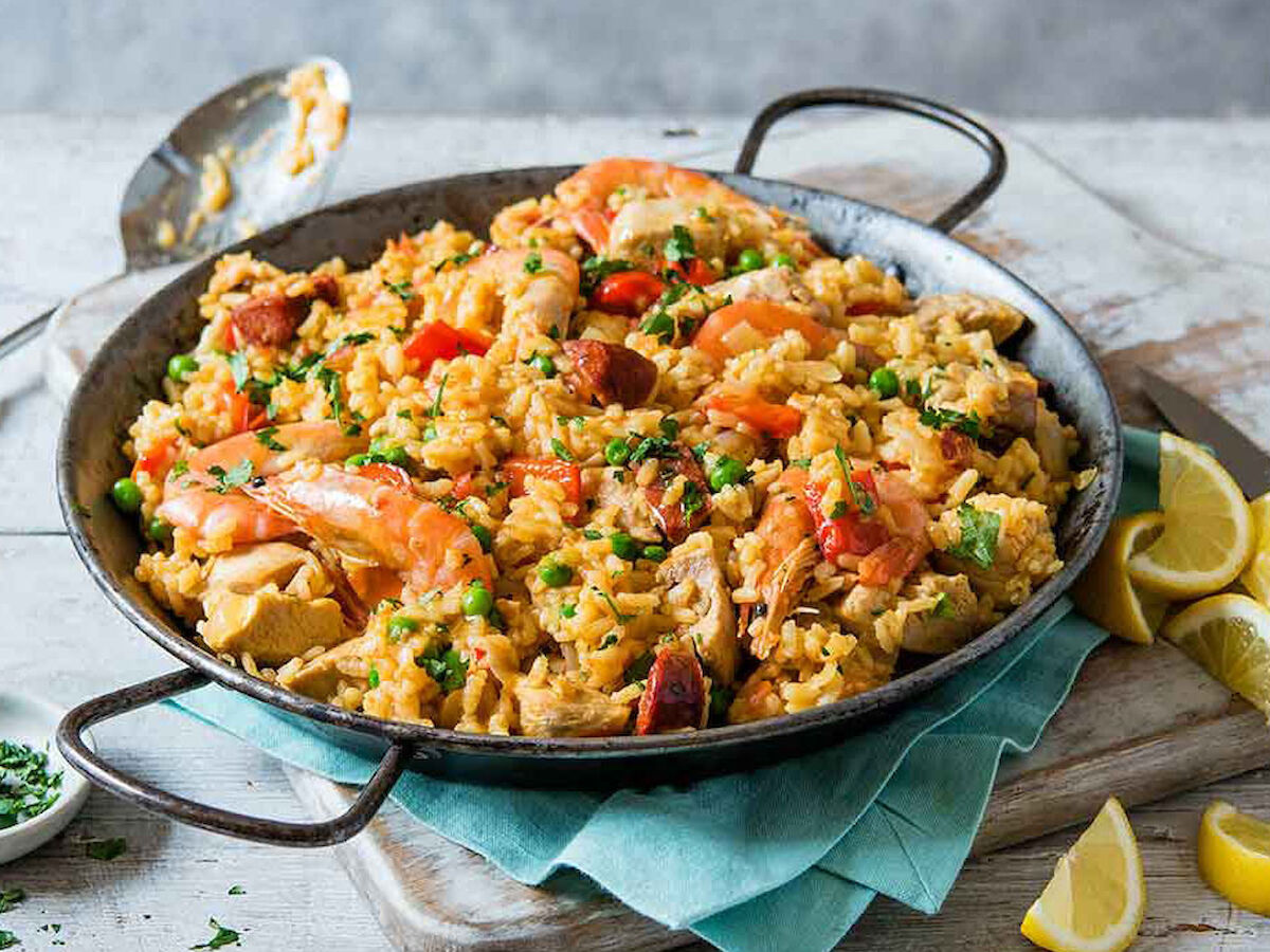 A pan of seafood paella with shrimp, mussels, rice, and vegetables garnished with chopped parsley, set on a cloth with lemon wedges on the side.