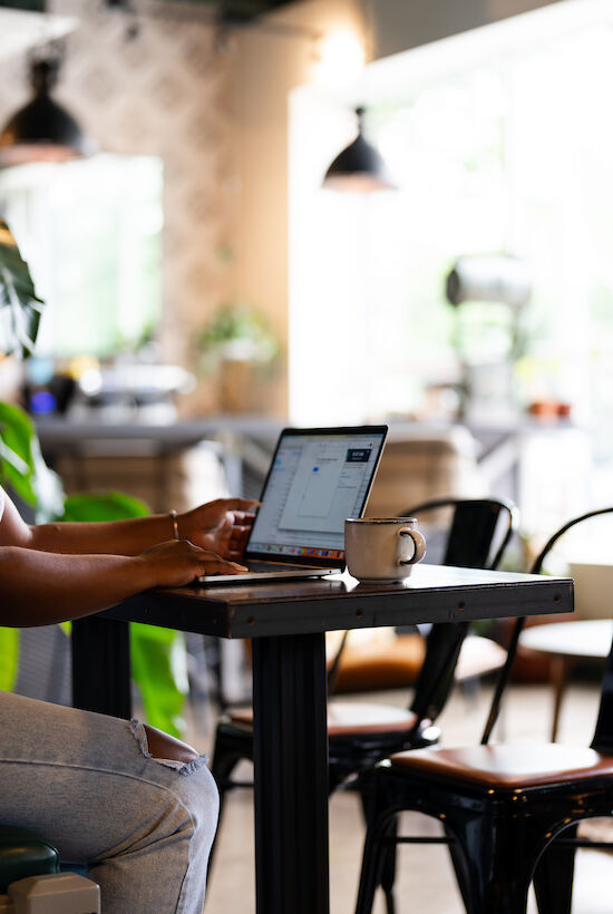 A woman is sitting at a table in a café, working on a laptop, with a coffee cup in front of her. The café interior is modern and cozy.