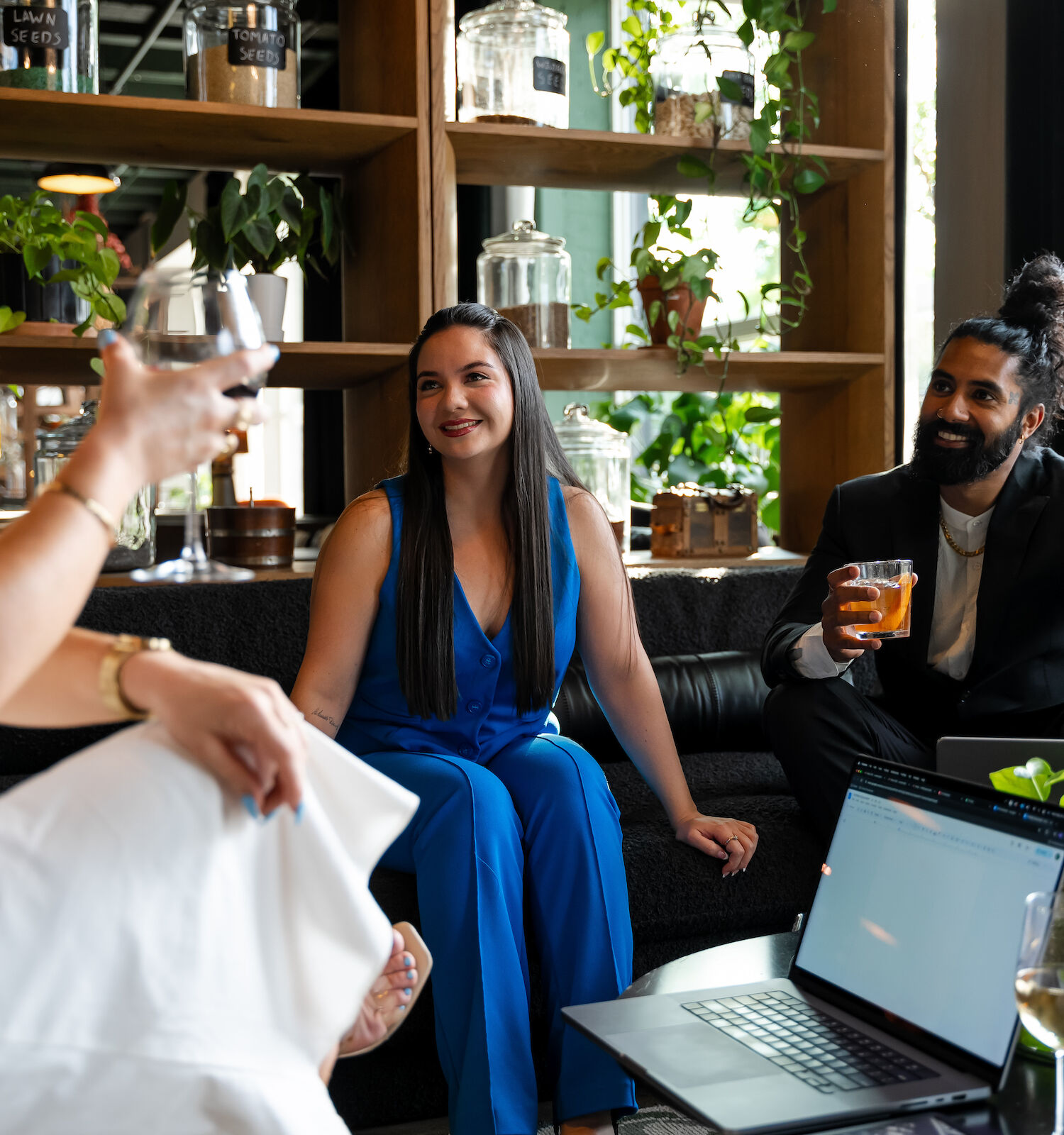 Three people in a social setting, talking and enjoying drinks with a laptop open on the table nearby and greenery in the background.