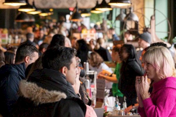 A crowd of people is gathered in a busy indoor market or food hall, engaging in conversations and transactions under hanging lights.