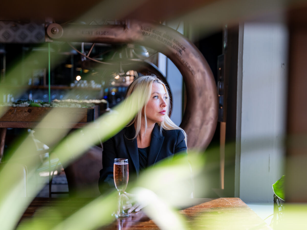 A woman is sitting at a table in a stylish indoor setting, with plants in the foreground. She has a glass of drink in front of her ending the sentence.