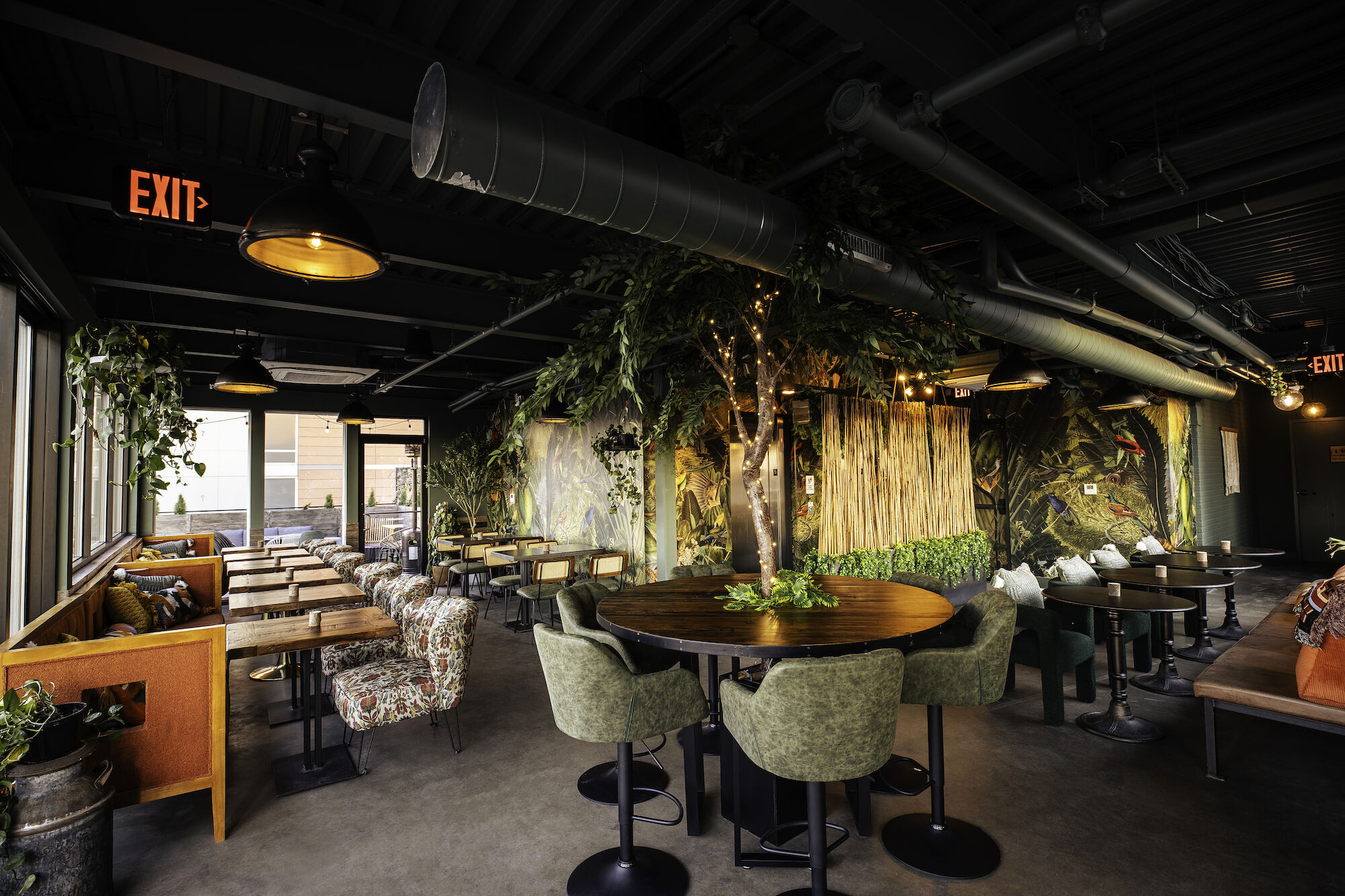 A cozy restaurant interior with green decor, plants, cushioned seating, wooden tables, and an exposed ceiling, creating a relaxing ambiance.