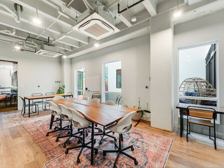 A modern conference room with a wooden table, chairs on wheels, a rug, and large windows in a bright space with exposed ceilings and lights.