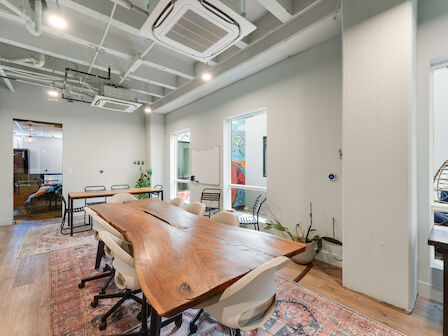 This image shows a modern conference room with a wooden table, several chairs, plants, and large windows. The room has a bright and clean aesthetic.