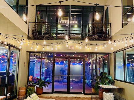 A cozy outdoor courtyard with string lights, modern seating, plants, and tables surrounded by a two-story building with large windows.