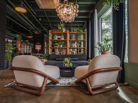 Cozy lounge area with comfortable chairs, a sofa, plants, a bookshelf, and a unique chandelier by large windows.