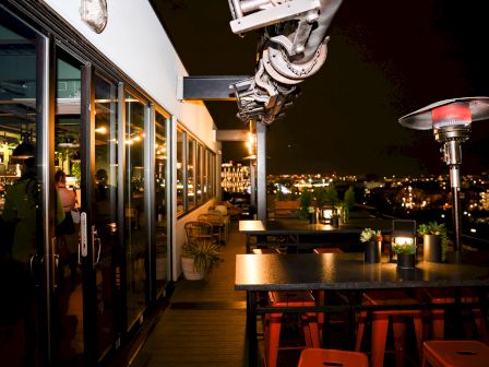 A rooftop bar at night featuring outdoor seating, a heating lamp, lush plants, and city views through glass doors leading to indoor seating, ending the sentence.