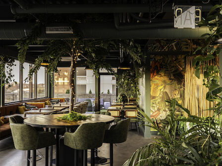 The image shows a modern dining area with green chairs, wooden tables, and abundant indoor plants, providing a lush and inviting ambiance.