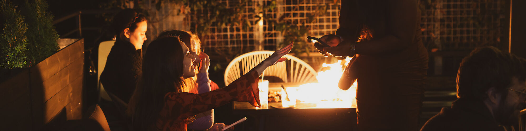 People sitting around a fire pit during what seems to be an evening gathering. Warm lighting illuminates the scene, with drinks on the table.