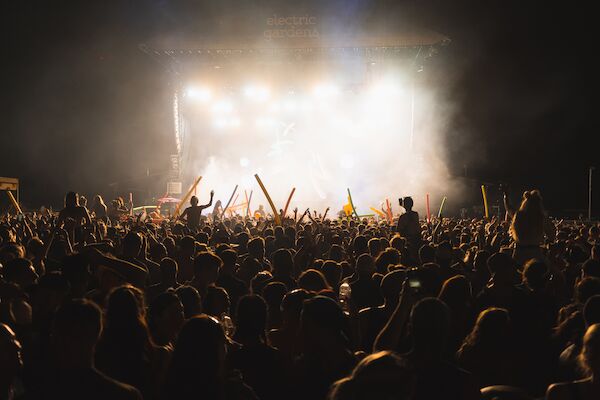 A large, energetic crowd is gathered at an outdoor concert at night, with people raising their hands and glow sticks under stage lights.