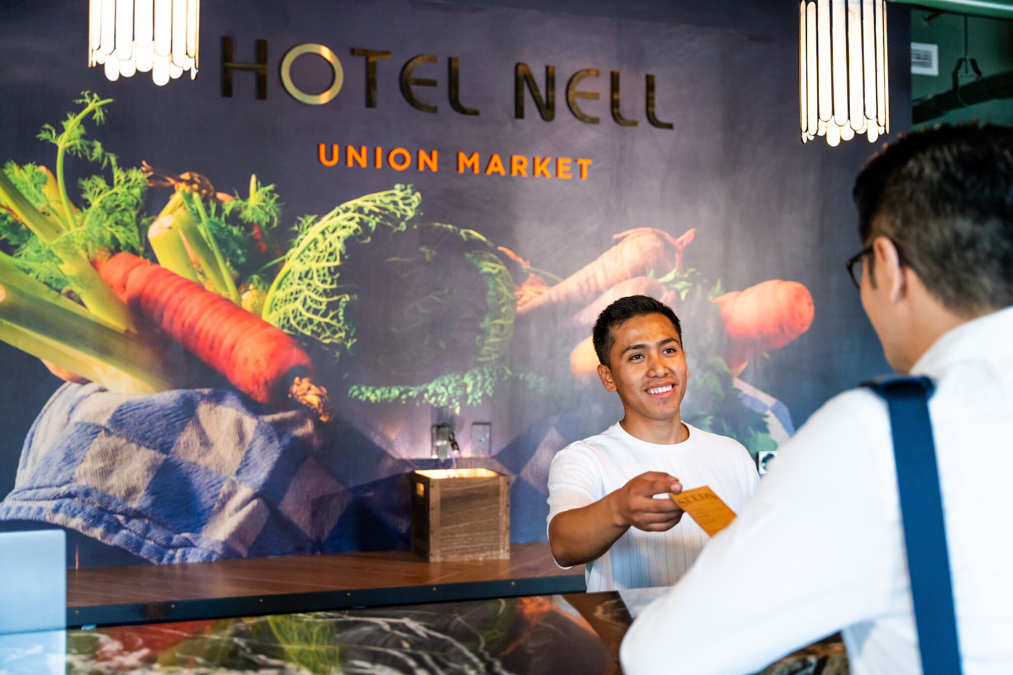 A front desk staff member at Hotel Nell's Union Market is handing a key card to a guest, with a colorful vegetable-themed background mural.