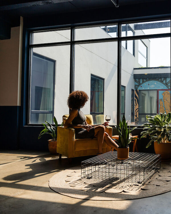 A person sits in a sunlit room with plants, looking out a large window at a mural that says 