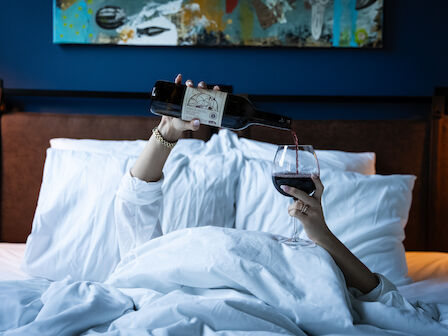 A person in bed pours red wine into a glass while surrounded by white linens and pillows, with abstract art on the wall behind them.