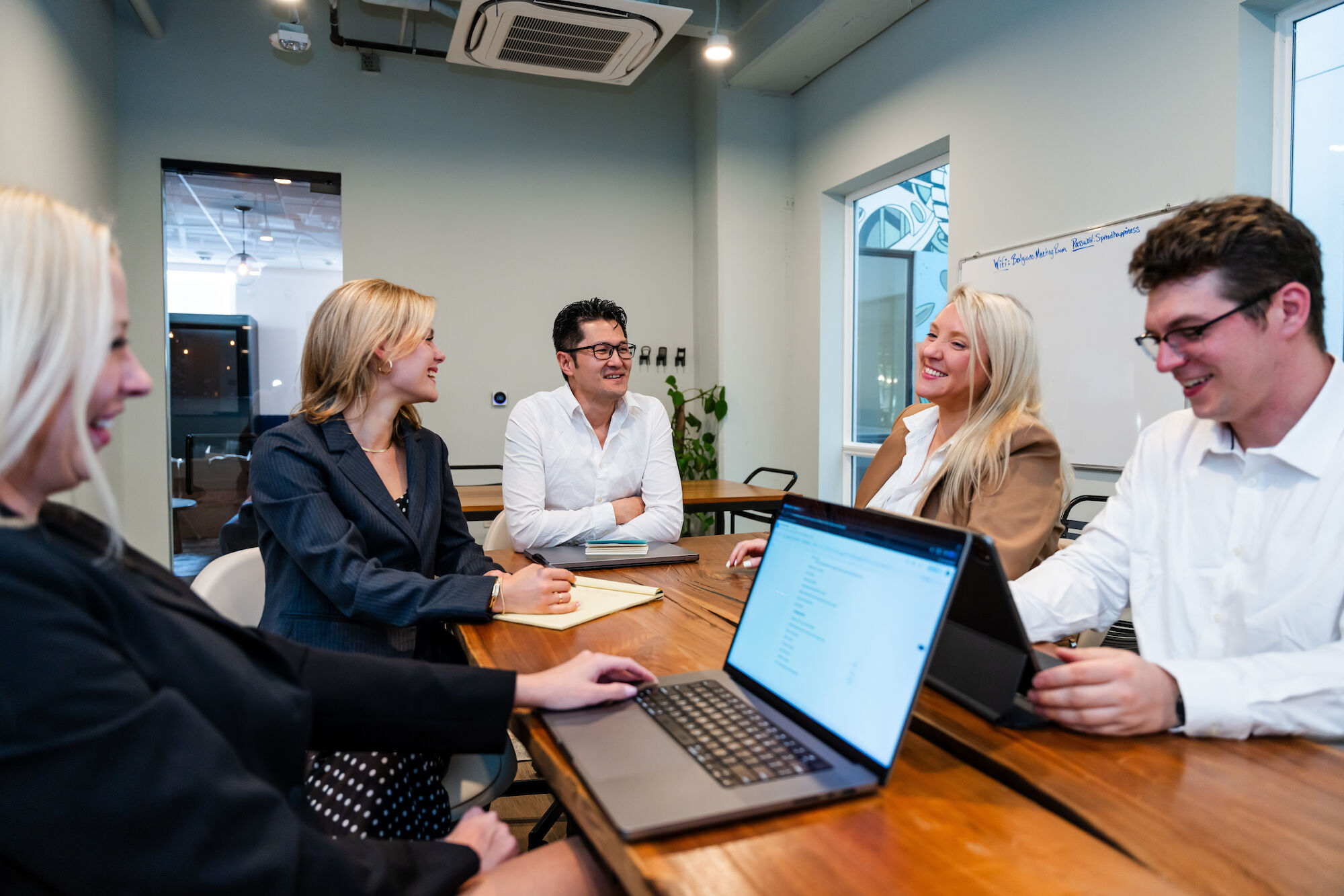 A group of people is engaged in a meeting around a table, with laptops and notebooks. They are smiling and appear to be having a discussion.