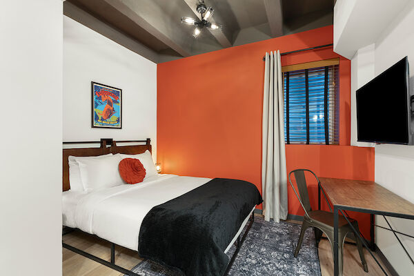 A cozy bedroom with an orange accent wall, a bed with white linens and a black throw, a desk with a chair, a wall-mounted TV, and a painting.