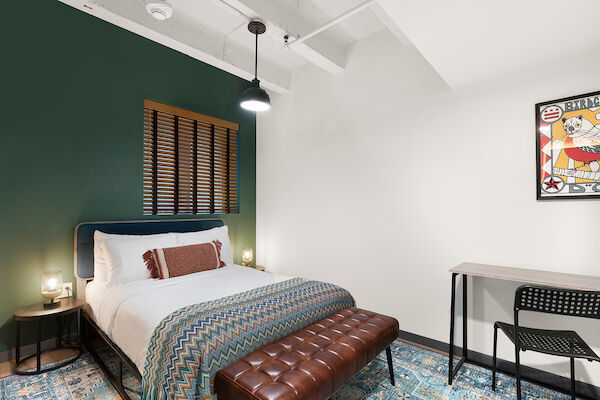 A well-decorated bedroom with a bed, a brown bench, a desk with a chair, a wall lamp, a window, and a colorful wall hanging.