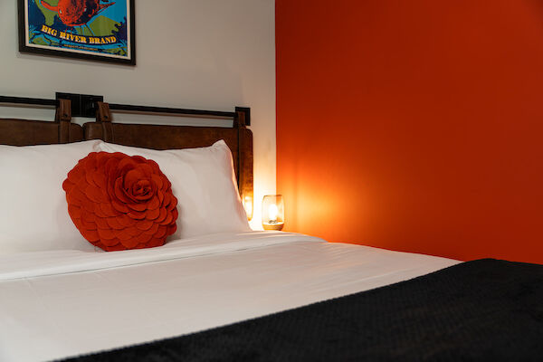 A well-made bed with white linens, a large orange flower pillow, an orange accent wall, a small lamp, and a framed poster on the wall.