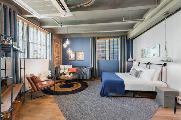 A modern, stylish bedroom with industrial elements, featuring a large bed, sitting area with chairs, coffee table, and blue accents throughout the room.