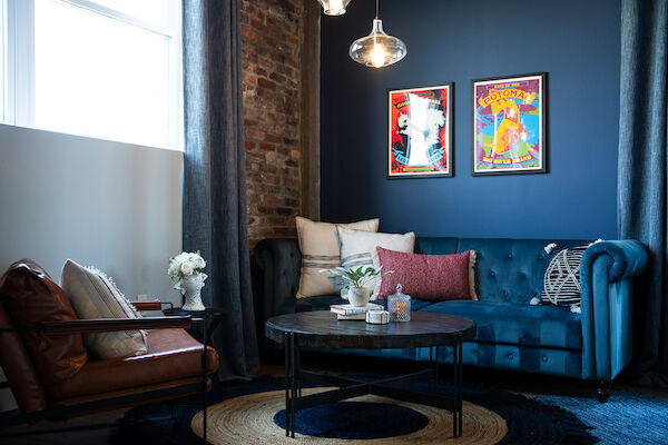 A cozy living room with a brown armchair, blue couch, round coffee table, two framed posters on a blue wall, and decorative plants.