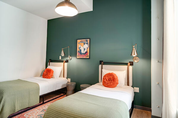 A bedroom with two single beds, each with a green blanket and orange cushion, green wall, wall lamps, art, and a pendant light.