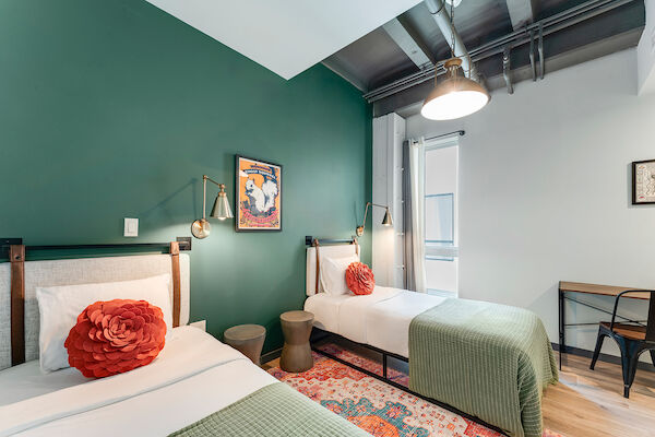 A cozy room with two single beds, green bedding, red pillows, a small desk, wall lamps, a rug, artwork, and an overhead light.