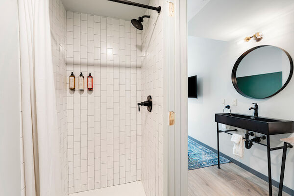 The image shows a modern bathroom with a tiled shower area, black fixtures, and a vanity with a round mirror and wall-mounted lights.