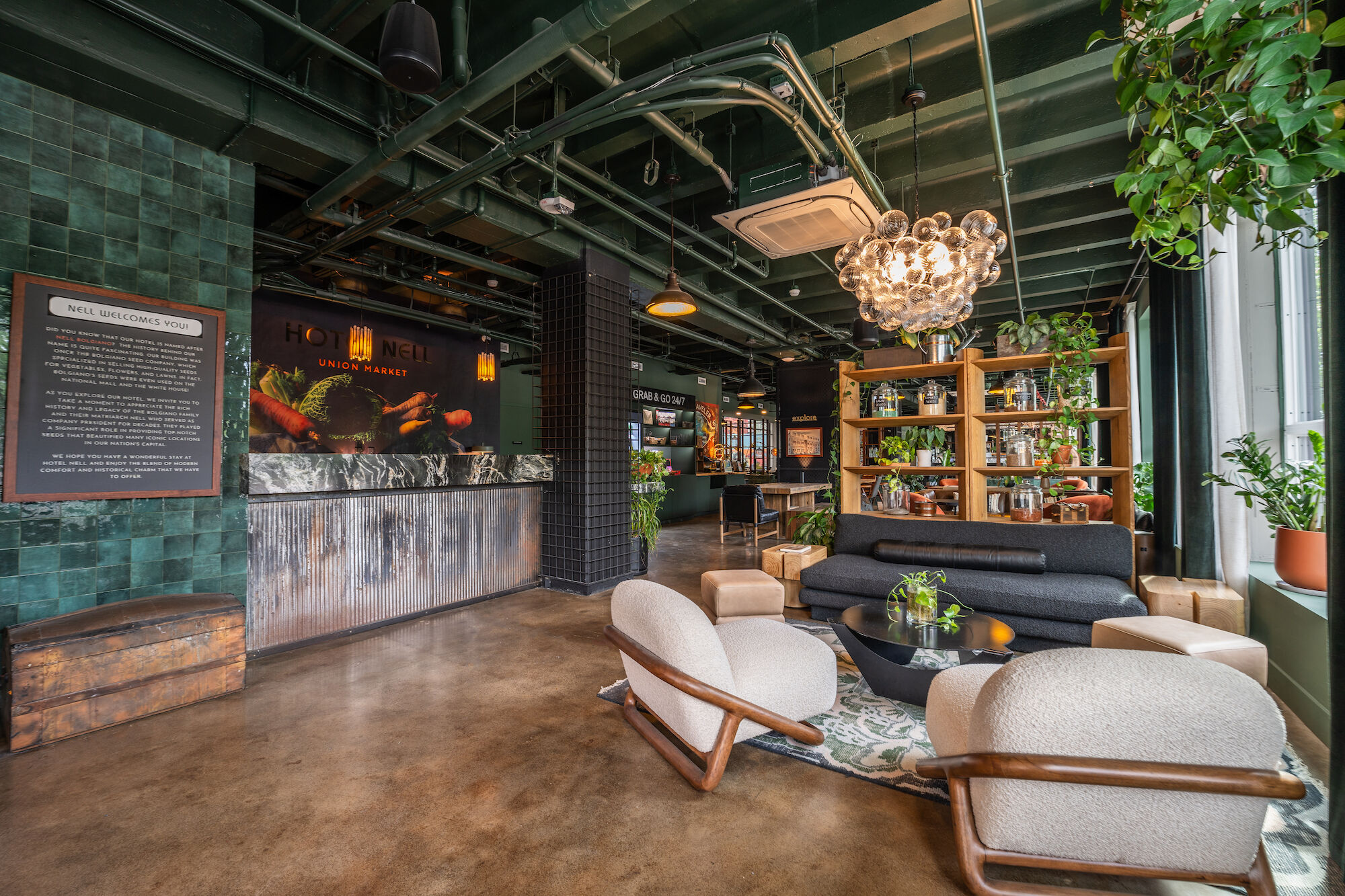 A modern lounge area with stylish seating, a chandelier, plants, and a reception desk. Decor includes bookshelves and green tiles ending the sentence.