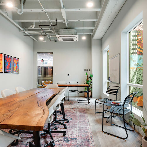 The image shows a modern conference room with a wooden table, various chairs, wall art, a whiteboard, and a large window allowing natural light.