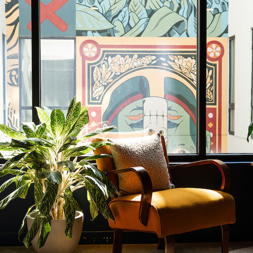 A cozy yellow chair with a cushion is placed next to a green potted plant, with a colorful mural on the building visible through the windows.