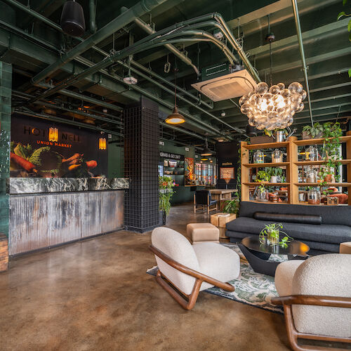 A modern lobby with green and beige decor, plants, a lounge area with chairs and a couch, a bookshelf, and a reception counter.