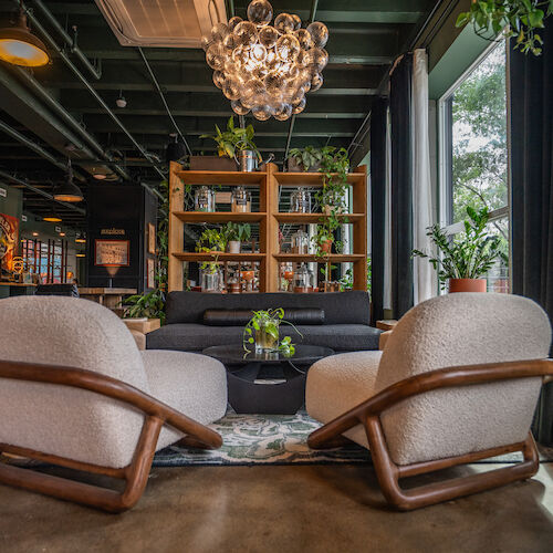 The image shows a stylish, green-themed seating area with modern furniture, a unique chandelier, and abundant plants and natural light from large windows.