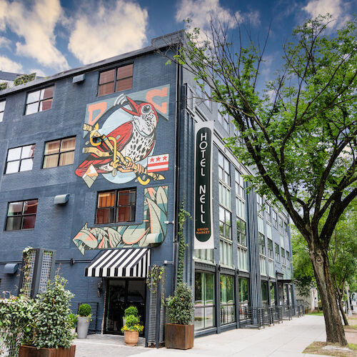 This image shows the exterior of Hotel Nelk with a mural on its wall, surrounded by greenery and a tree-lined street under a partly cloudy sky.