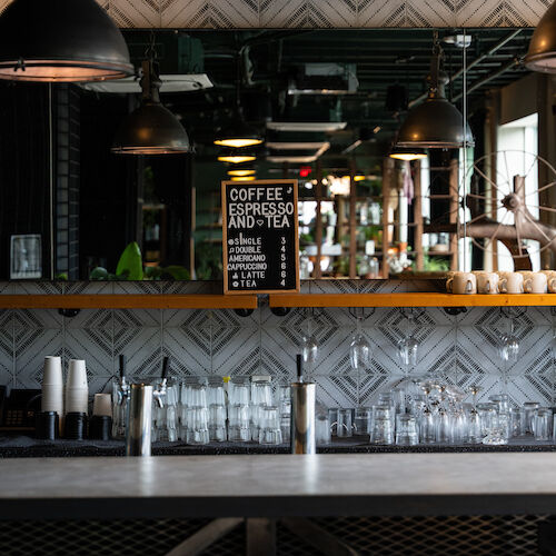 A modern bar interior with hanging lights, a mirror, bar stools, and various bottles and glasses on the counter.