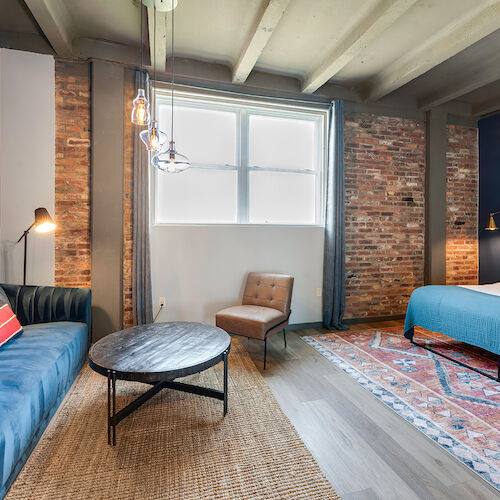 A cozy room features a bed, blue sofa, rug, chair, coffee table, hanging lights, brick walls, and a large window with frosted glass.